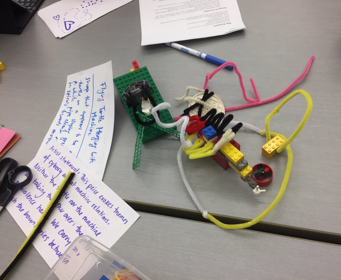 An image showing a small sculpture made of lego and pipe cleaners on
a table top, beside which lie two hand-written notes, one of which includes a title and description, while the other
contains an artist statement.