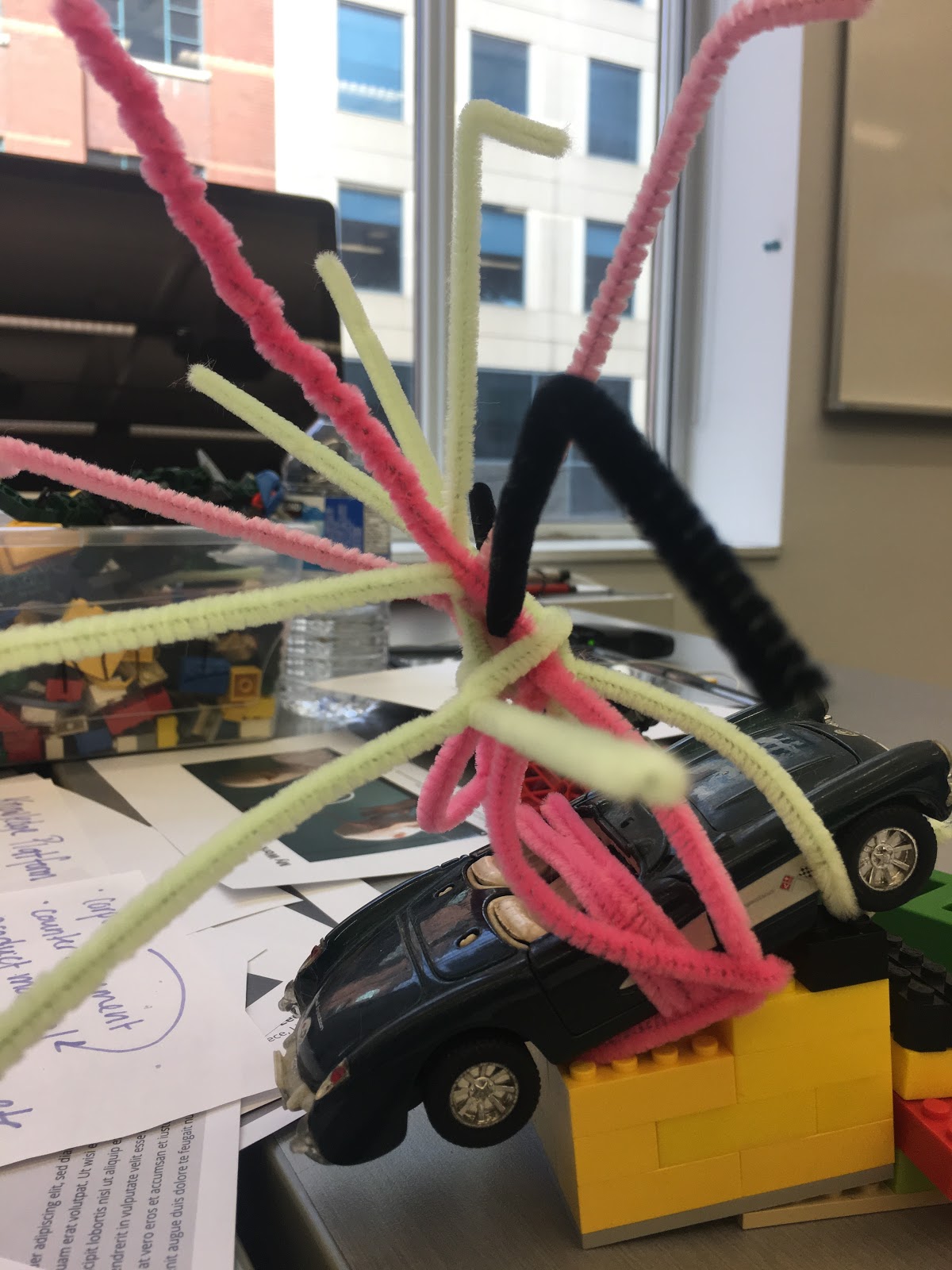 La photographie d'une sculpture faite à partir de légos d'une petite
voiture et de fil chenille. Sur la même table, on peut voir des notes écrites à la main ainsi qu'une boîte contenant des
briques de couleur en arrière plan.