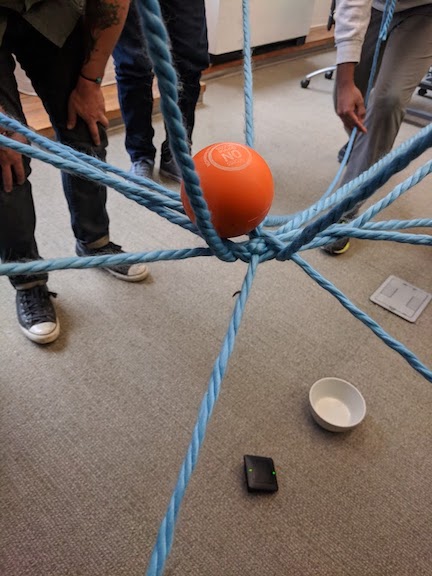 An image showing a closeup view of the intersection point of the
web of strings raised above the floor, on which balances an orange ball. On the floor below a white bowl can be seen, as
well as several participants standing in the background holding the ends of the strings.
